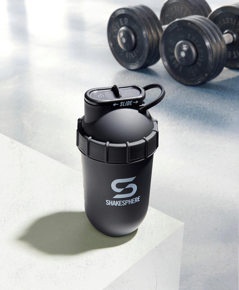 Ce shaker fitness utilise uniquement la force centrifuge créée par la forme arrondie de la capsule – sans aucun recoin dans lequel des résidus de poudre pourraient se coincer. Shaker noir sur une surface blanche avec des haltères en arrière-plan.