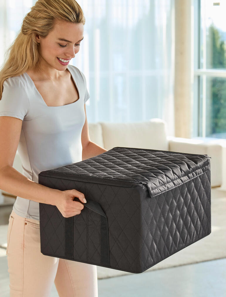 Des poignées robustes permettent un transport confortable et sûr. Femme soulevant une boîte empilable noire avec motif en losange, dans le salon.