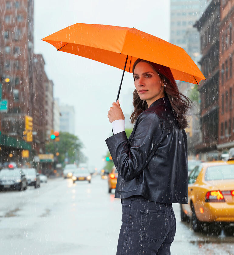 Femme tenant un parapluie orange en pluie, debout dans la rue.