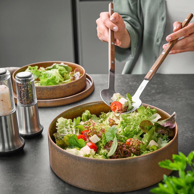 Personne tenant des couverts à salade XL au-dessus d'un bol de salade.