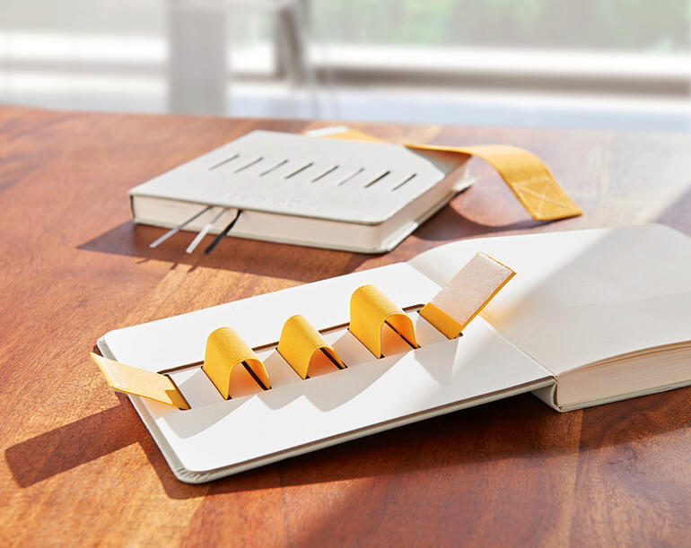 Le support élastique maintient stylos, smartphone, lunettes de lecture et bien plus encore bien en place, sans glisser. Flex-Book posé sur une table avec des bandes jaunes et un autre livre.