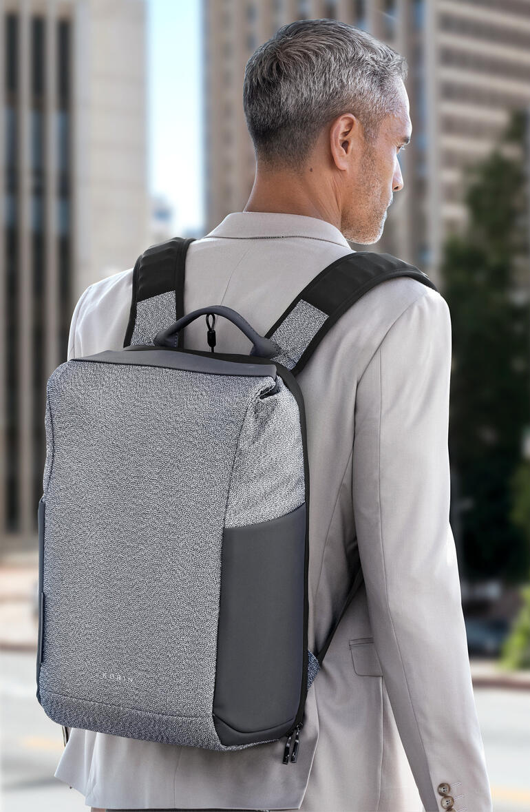 Homme avec un sac à dos antivol gris sur le dos devant un paysage urbain.