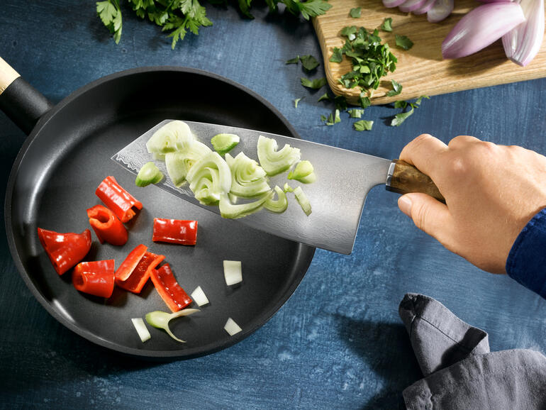 Sur la large lame, vous transférez vos aliments découpés proprement dans la poêle ou la casserole. Main tenant un couteau Nakiri en damas coupant des légumes dans une poêle.