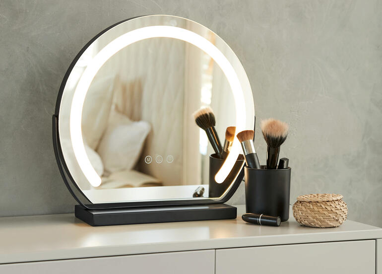 Miroir de maquillage rond avec éclairage sur une table, entouré d'ustensiles de maquillage.