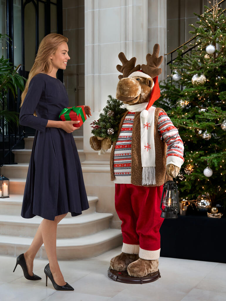 Élan dansant en tenue festive, tenant un cadeau devant un sapin de Noël.