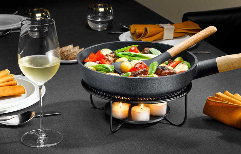 Poêle avec légumes sur un réchaud, à côté d'un verre de vin sur une table dressée.