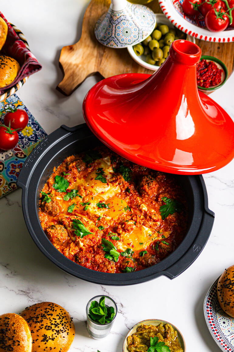 Tajine Peugeot Saveurs avec couvercle rouge, rempli de légumes colorés et d'herbes, sur une table avec des accompagnements.