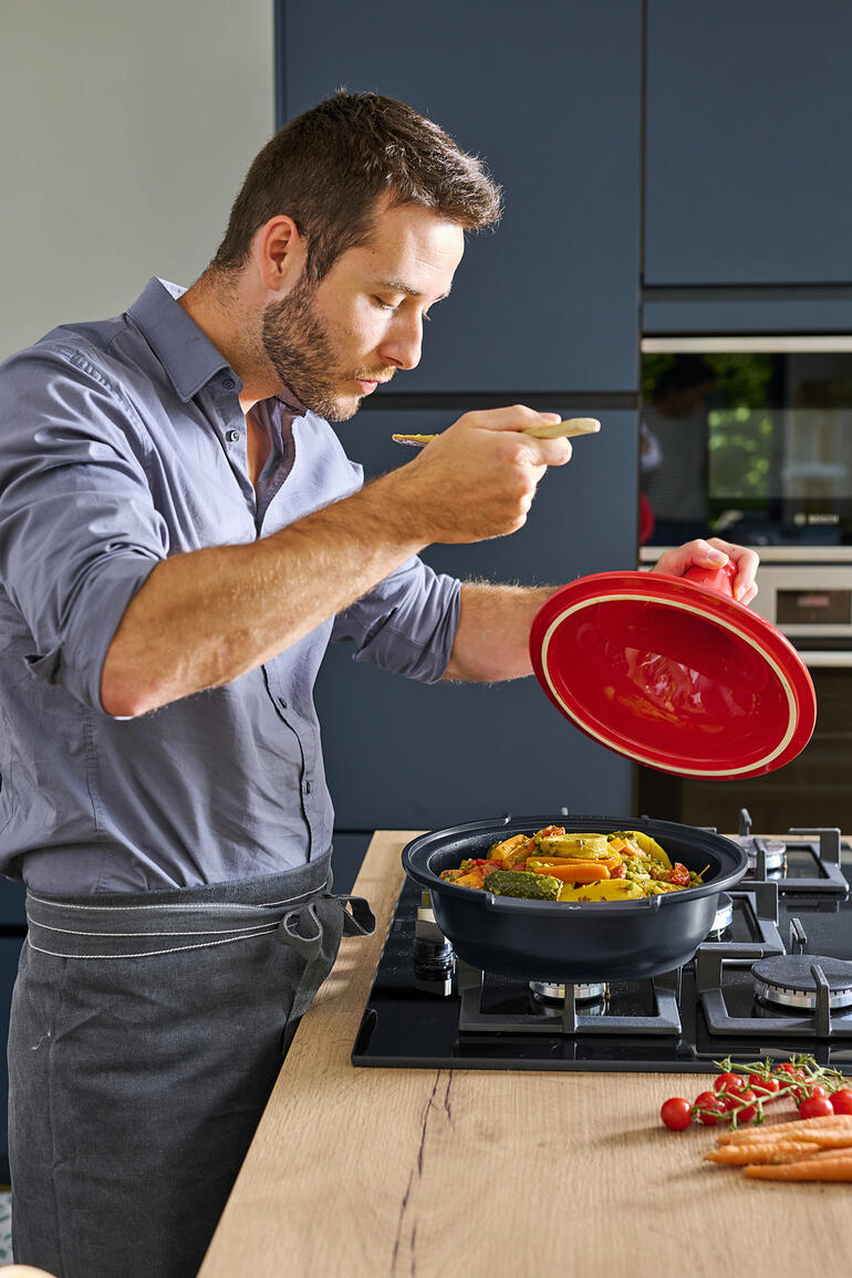 Homme dans la cuisine goûtant la tajine Peugeot Saveurs avec une fourchette tout en soulevant le couvercle.