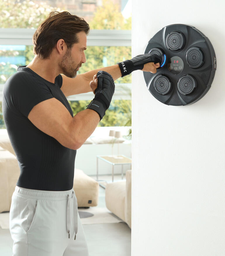Homme frappe avec ses mains contre un boxeur mural, salle de fitness avec fenêtres lumineuses en arrière-plan.