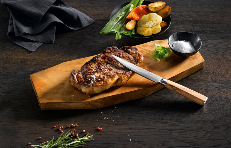 Couteau à steak en damas sur une planche en bois avec un steak grillé et des légumes.