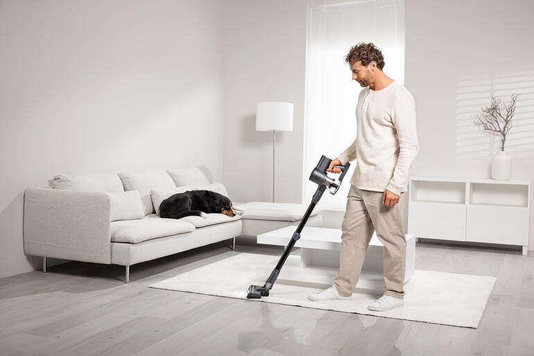 Un homme passe l'aspirateur sans fil sur un tapis, un chien est allongé sur un canapé.