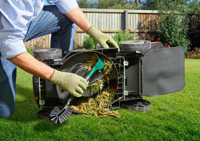 Personne nettoie une tondeuse à gazon retournée avec un outil, en enlevant les résidus d'herbe.