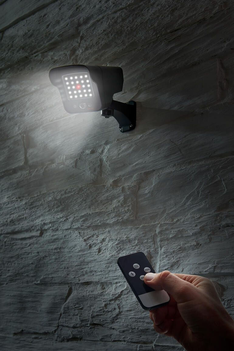 Personne avec télécommande activant une imitation de caméra solaire fixée sur un mur gris.