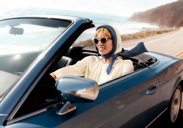 Femme dans une décapotable avec un foulard et des lunettes de soleil conduisant le long de la côte.