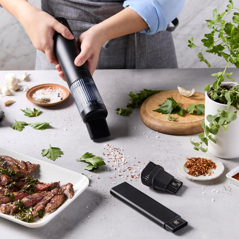 Aspirateur à main sans fil XTEND tenu au-dessus d'un morceau de viande, avec des herbes et des épices fraîches à côté.