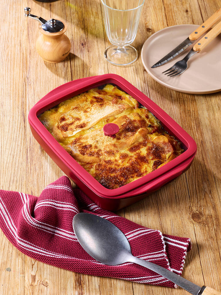 Plat à four rouge avec couvercle, rempli d'un plat de haricots, sur une table en bois.