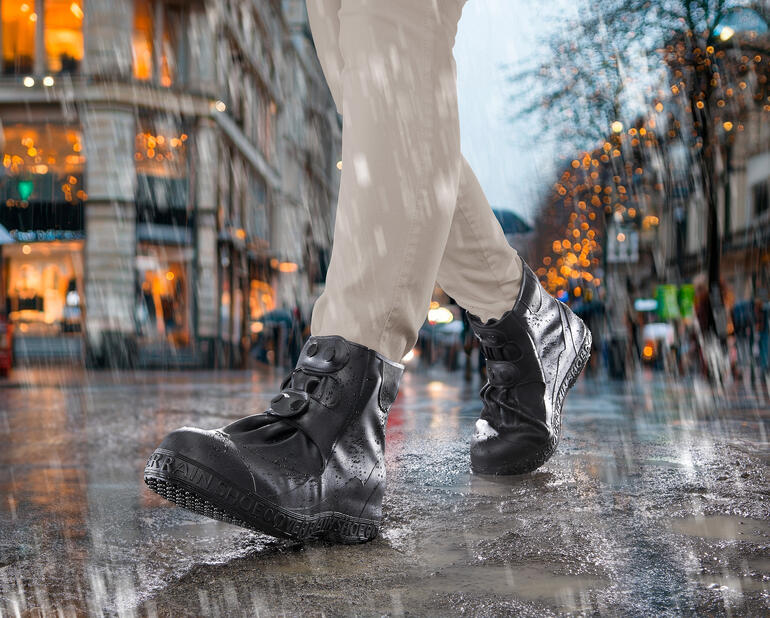 Personne portant des bottes noires avec des couvre-chaussures en silicone sous la pluie sur une rue mouillée.
