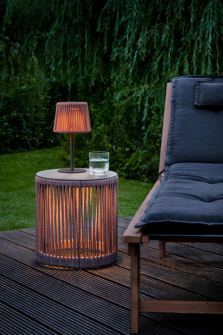 Table de jardin LED avec lampe orange et verre d'eau sur le terrasse en bois à côté d'un fauteuil.