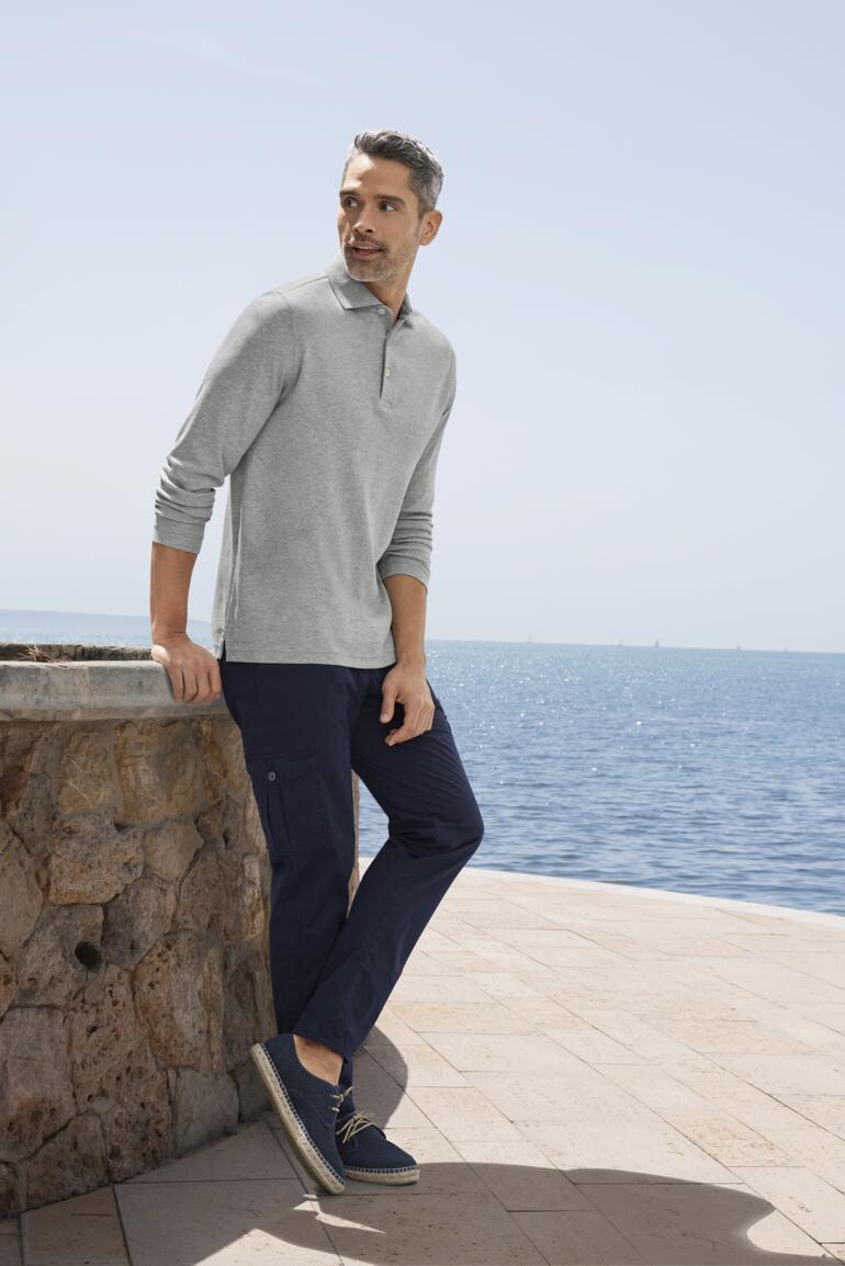 Homme portant un polo Pima interlock gris, debout près d'un mur en pierre avec vue sur la mer.