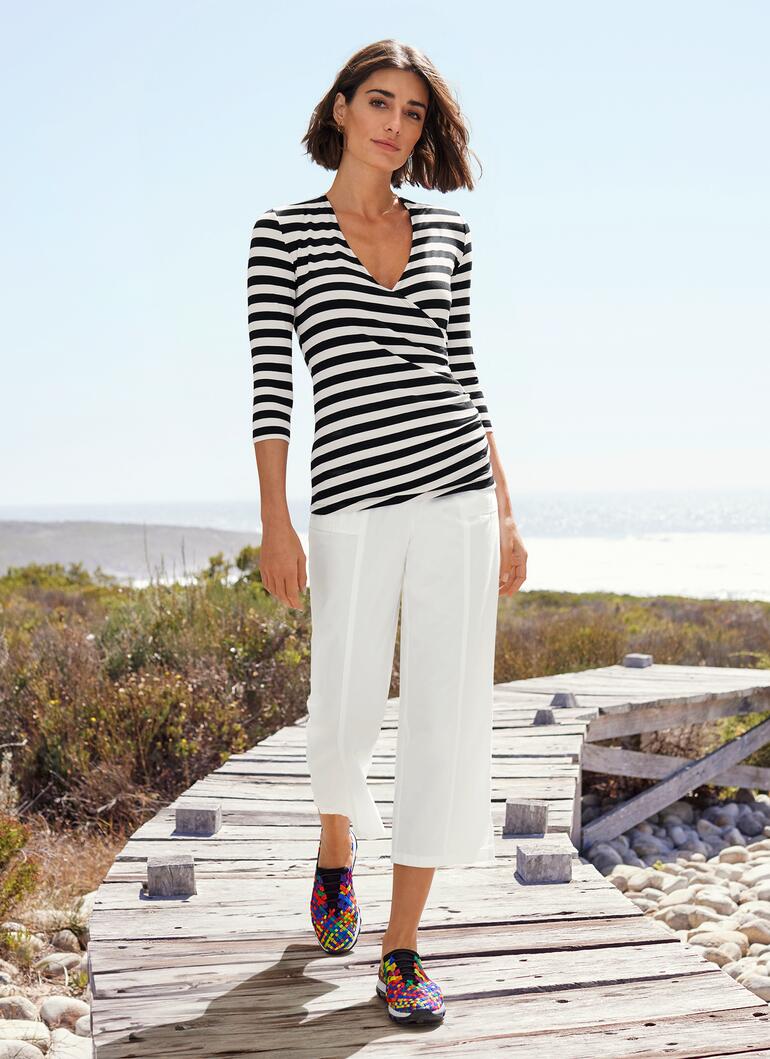 Femme en blouse rayée et pantalon blanc, sur un pont en bois.