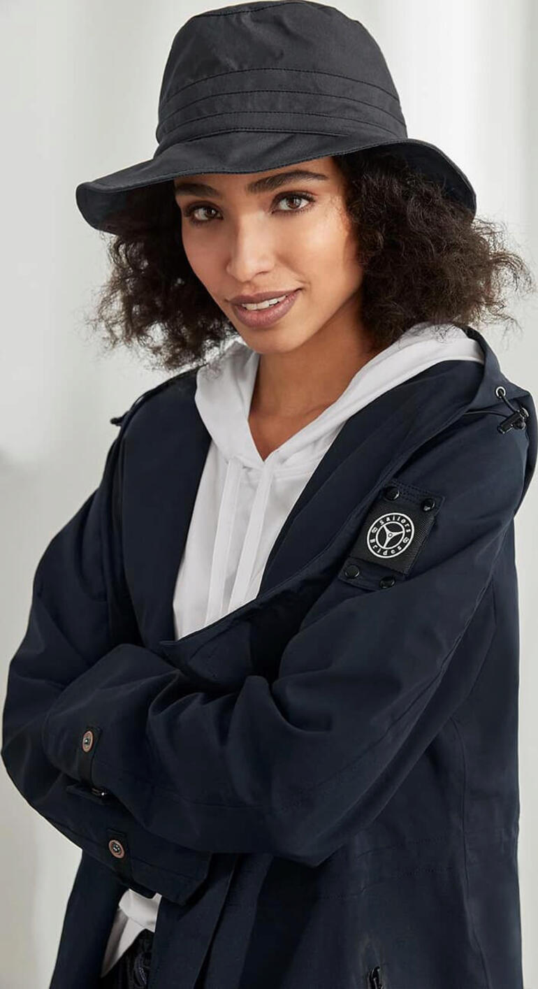 Femme portant un chapeau en coton noir pour tous les temps et un manteau gris foncé sourit à l'appareil photo.