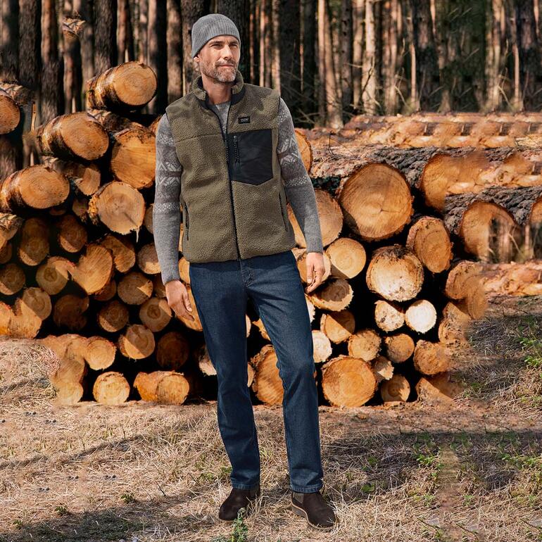 Gris Homme portant un pull norvégien en bébé alpaga, devant une pile de bois dans la forêt.