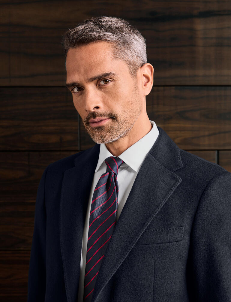 Homme avec cravate en soie Ascot, regard sceptique, devant un fond en bois sombre.