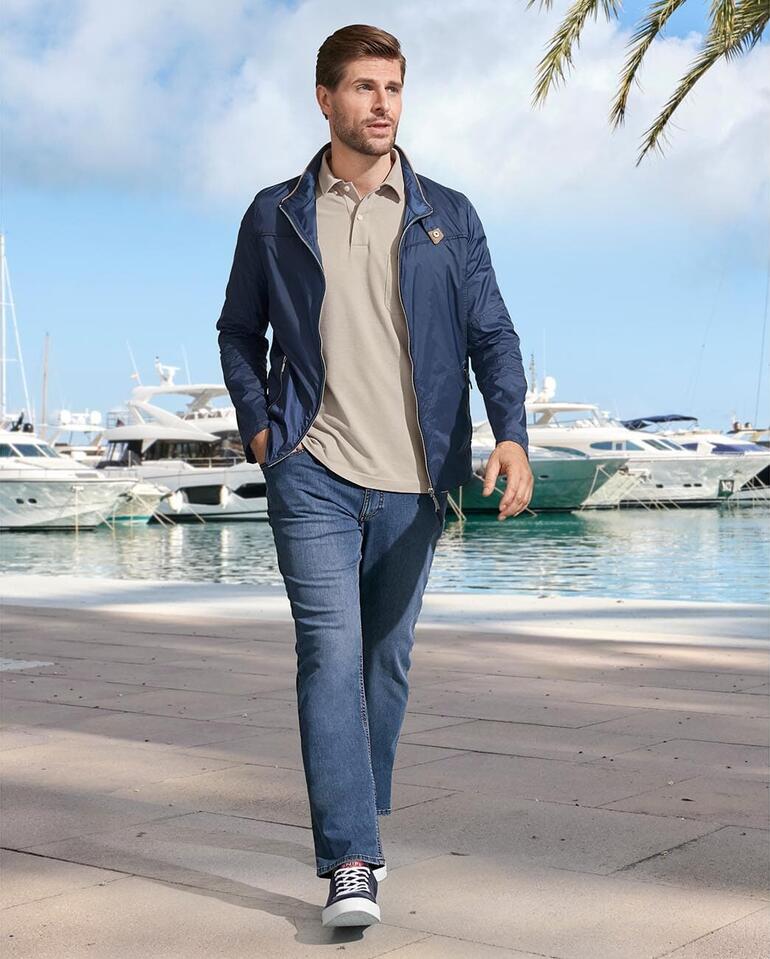 Jeans d’été Eurex by Brax Homme en veste bleue et polo beige marchant le long du port.