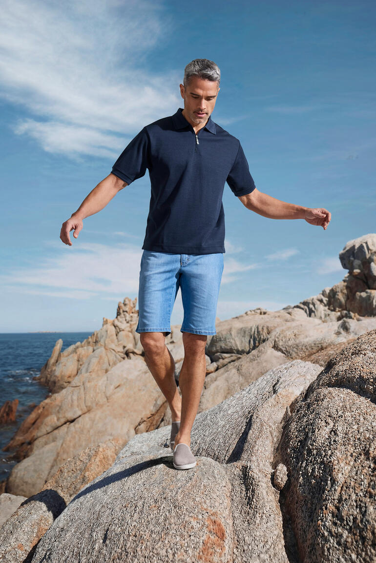 Homme portant un polo Coolmax à manches courtes marine, debout sur des rochers au bord de la mer.