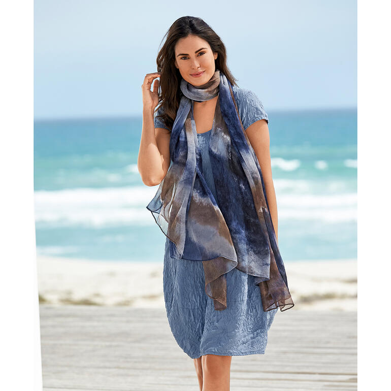 Femme en robe d'été en soie bleue marchant sur la plage avec une écharpe.