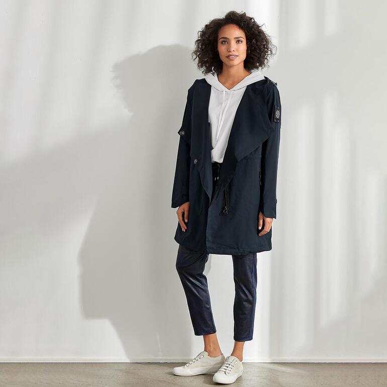 Femme dans un manteau softshell avec capuche, debout dans une pièce lumineuse.