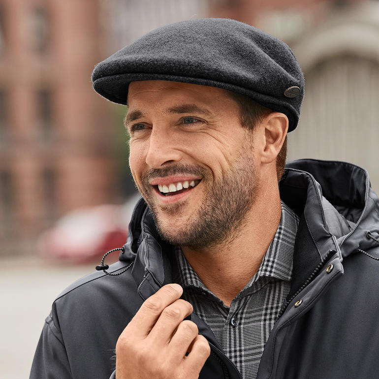 Homme avec une casquette Kangol et des protège-oreilles, souriant dans un environnement urbain.