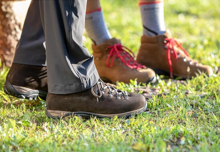 Homme, Marron foncé et Femme, Brun Bottes de randonnée Zamberlan®, pieds d'une personne dans des chaussures brunes et beiges sur une pelouse.
