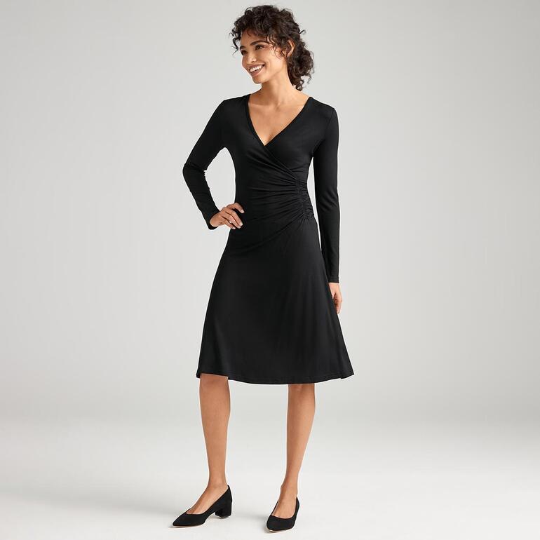 Femme en robe noire à manches longues, élégamment debout dans une pièce claire.