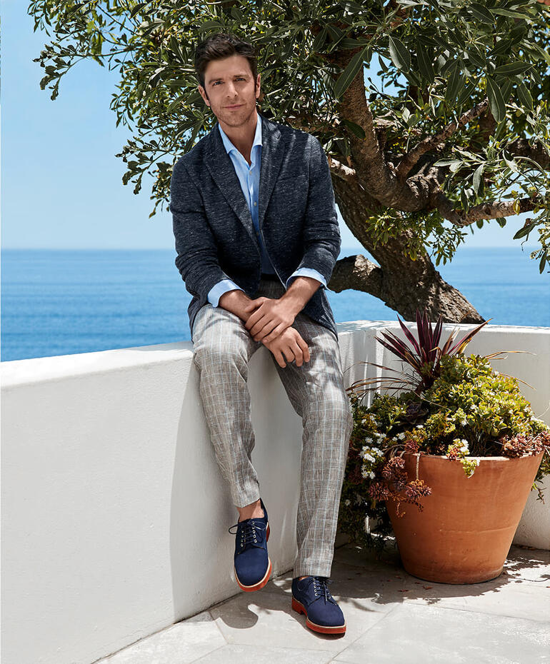 Pantalon en lin prince de galles de Bottoli Homme en blazer en jersey de lin Barutti, assis sur un mur près de la mer.