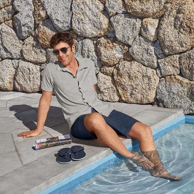 Marine Un homme assis au bord de la piscine dans une chemise à manches courtes grise et des lunettes de soleil.