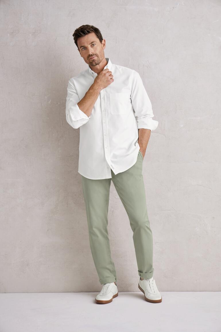 Sauge Homme en chemise blanche et chinos verts se tient devant un mur gris.