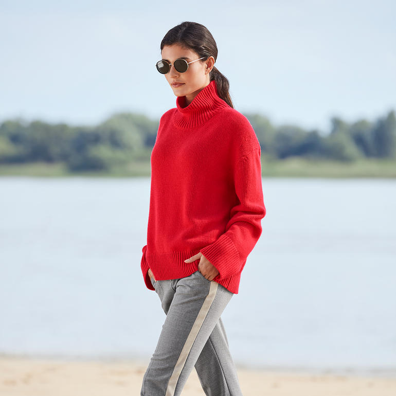 Femme portant un pull rouge avec bandes latérales, marchant élégamment au bord de l'eau.