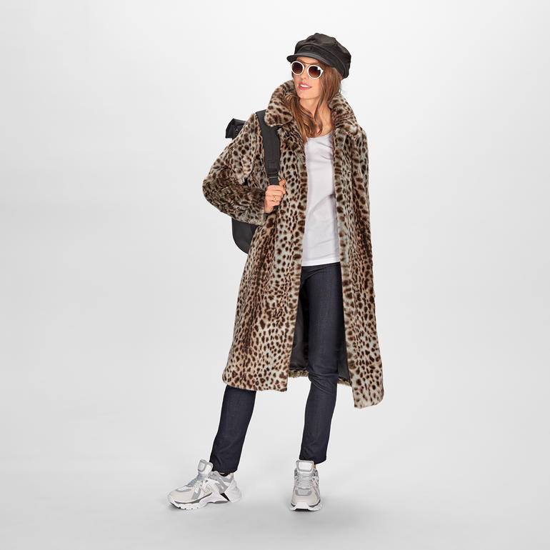Femme portant un manteau léopard avec des lunettes de soleil et un sac à dos sur fond blanc.