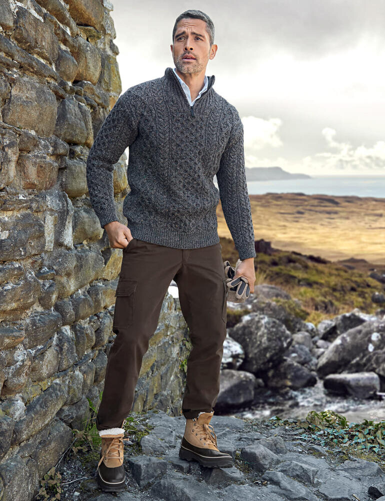 Pull Aran Irelands Eye Homme dans un pull Aran gris devant un mur de pierre dans la nature.