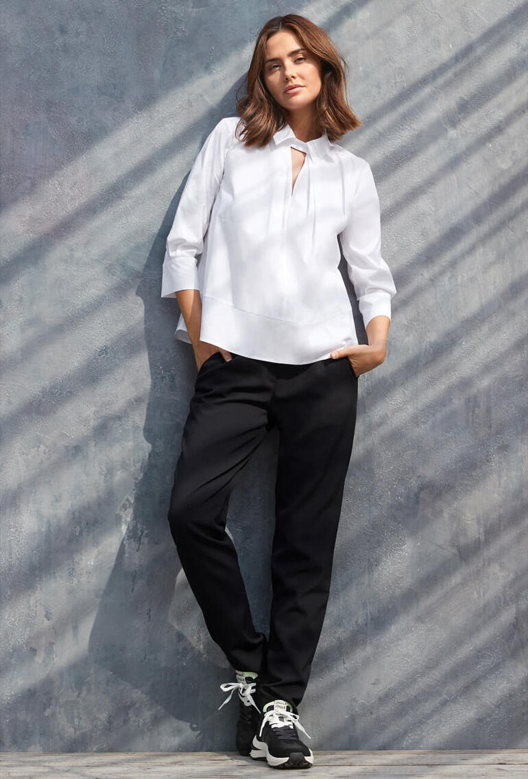 Femme en blouse blanche et pantalon noir se tenant contre un mur gris