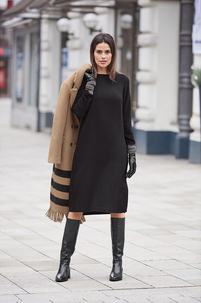 Femme en robe noire et manteau, avec des bottes longues sur une place.