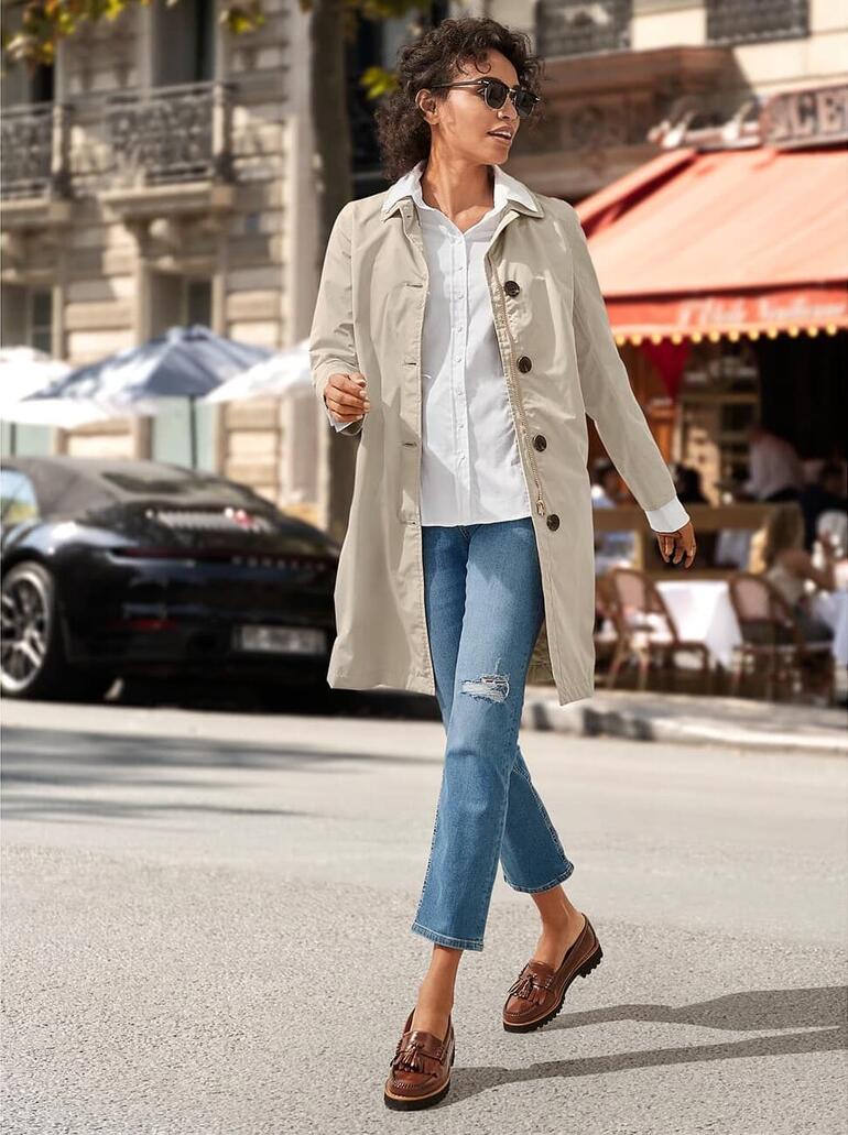 Femme en trench-coat beige et baskets blanches marchant dans une rue animée avec des cafés.