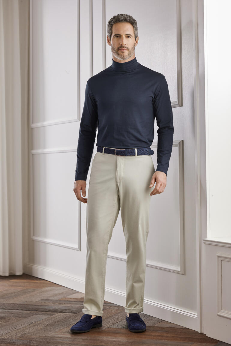 Crème Homme dans un sous-pull Sunspel sombre et un pantalon clair dans une pièce élégante.