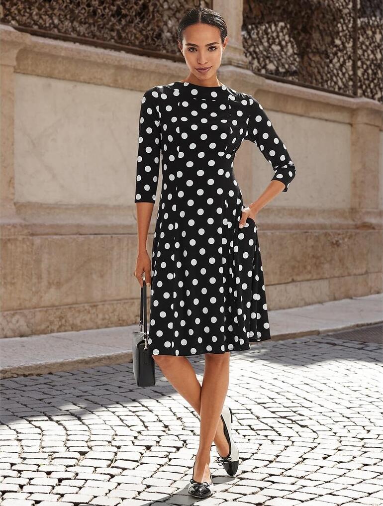 Femme en robe à pois se tenant dans la rue, souriante.