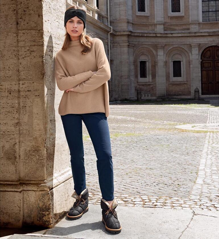 Femme en pull beige et jeans bleu se tient avec des chaussons en duvet Flufié sur un sol en pierre.