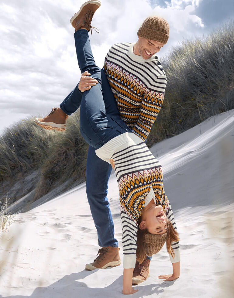 Homme et Femme Homme soulevant joyeusement une femme au-dessus de la plage, tous deux en pulls en jacquard.