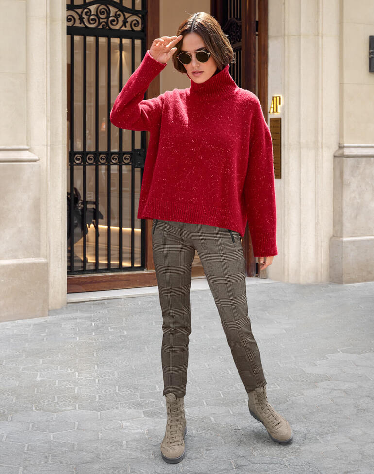 Femme en pull rouge et pantalon à carreaux devant une façade de bâtiment.