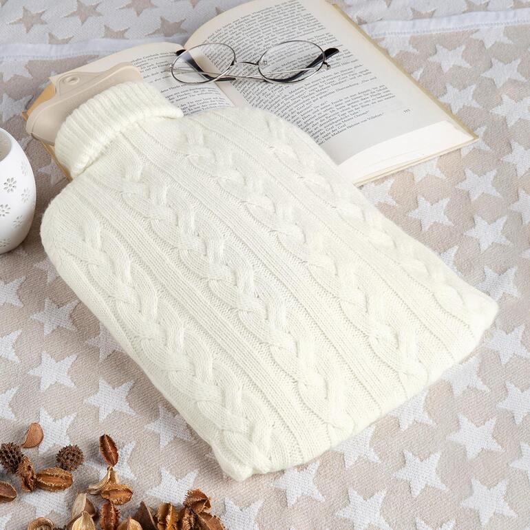 Bouillotte en cachemire dans une housse tricotée crème sur une table avec une page de livre et des lunettes.