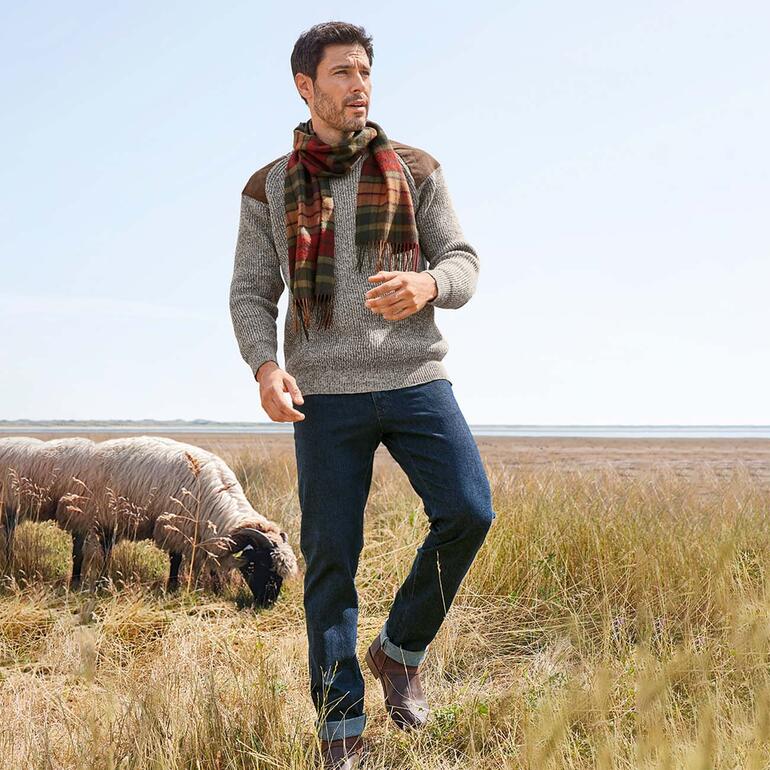 Homme en pull gris et écharpe à carreaux, debout dans un champ avec un mouton en arrière-plan.